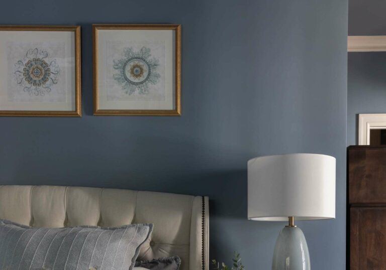 Revelry interior design image of a moody blue bedroom with 2 gold frames above an upholstered bed with a dark wood nightstand and a pale blue table lamp with gold picture frames